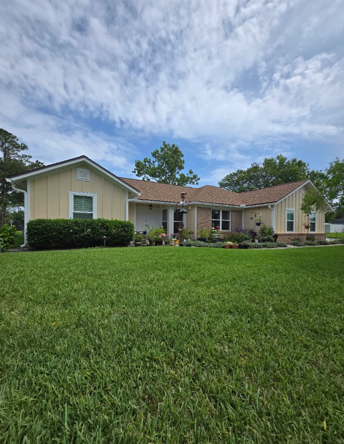 Single-story ranch home with pale yellow vertical board-and-batten siding, white window trim, and brick base