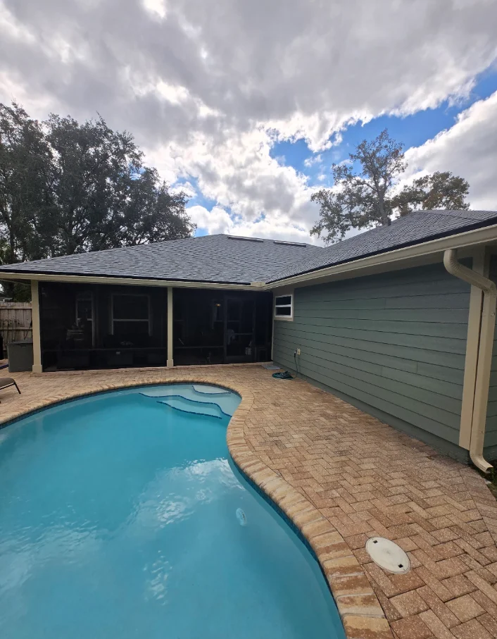Gray-green horizontal lap siding with cream trim and gutters on a home beside a kidney-shaped pool and brick paver deck