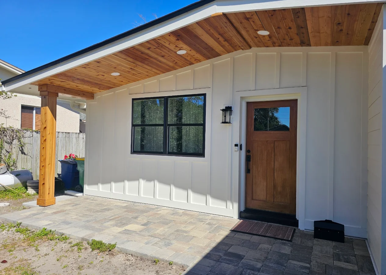 White board-and-batten siding with black-framed window, wood entry door, cedar porch ceiling, and paver walkway