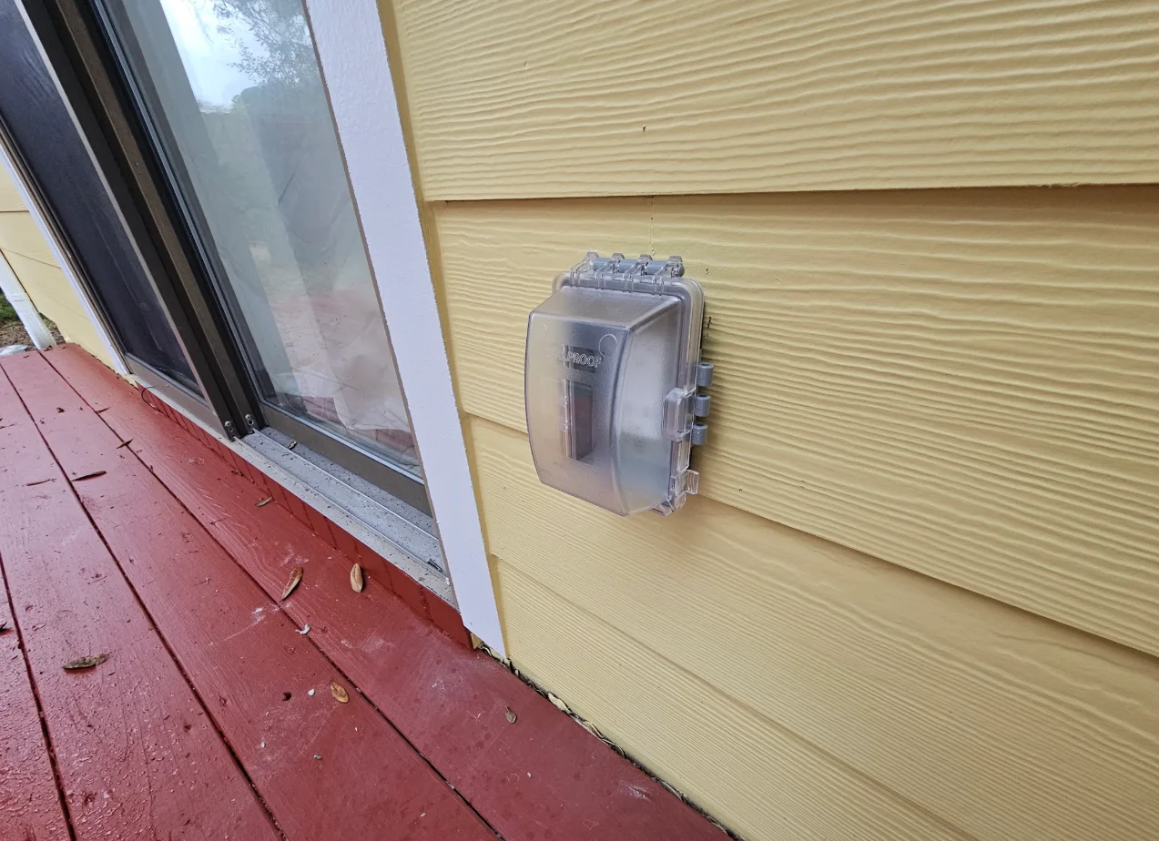 Clear weatherproof outlet cover mounted on yellow vinyl lap siding next to a white trim sliding door