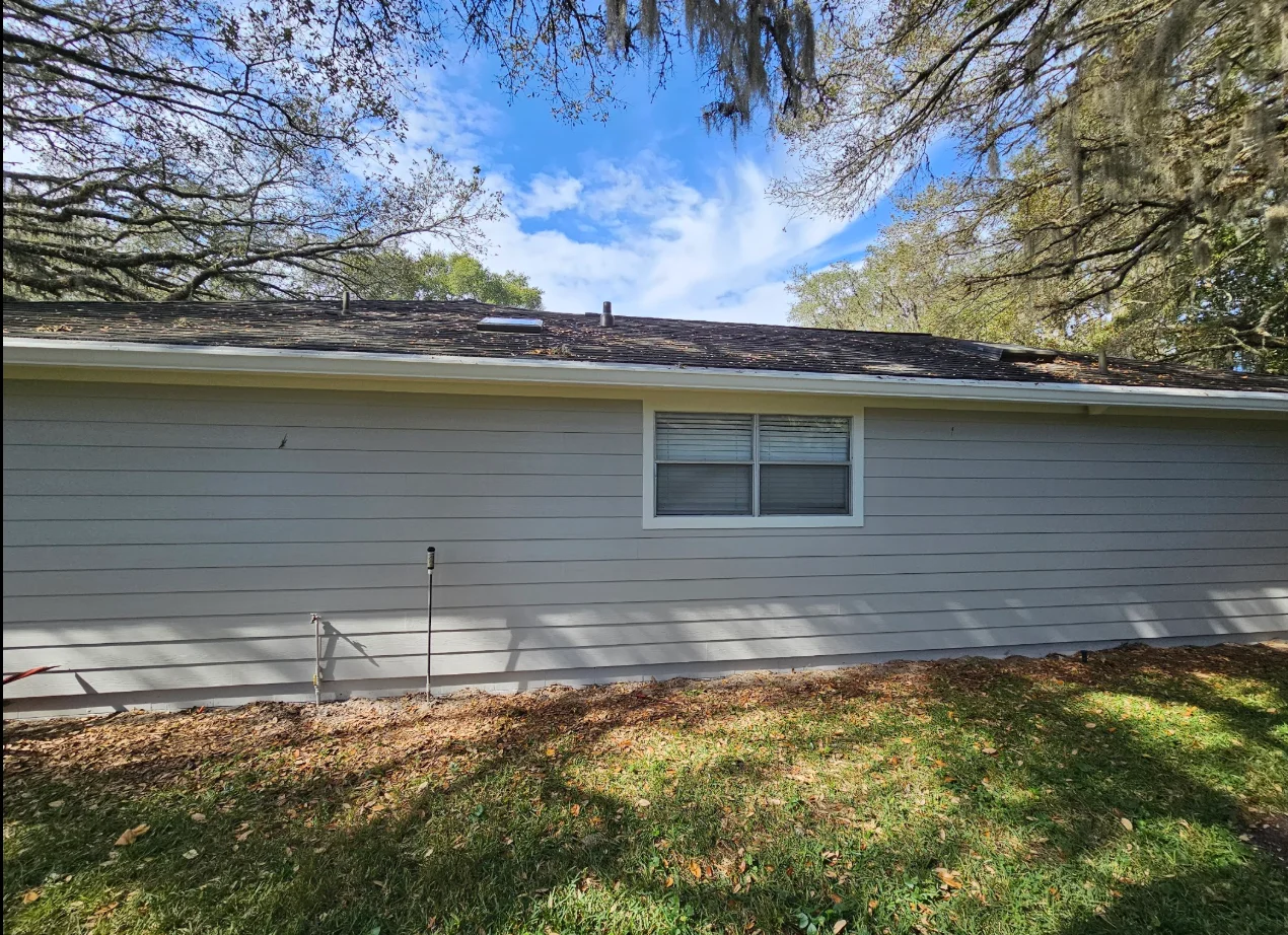 Gray horizontal lap siding with white window trim and gutter on a single-story home beneath oak trees