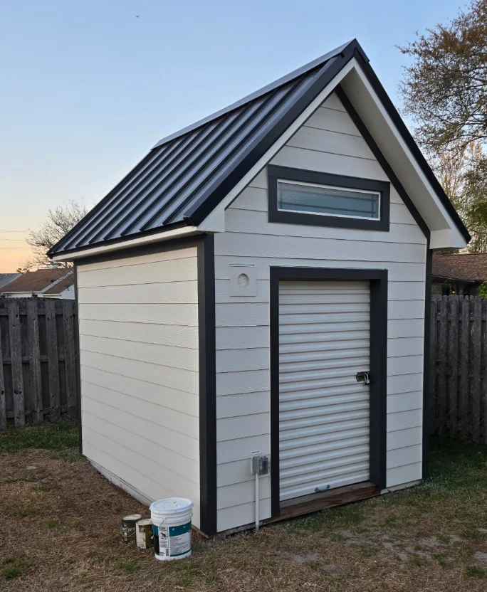 White horizontal lap siding and charcoal trim on a gable shed with standing-seam metal roof and roll-up door